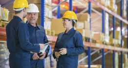 Drei Erwachsene mit Schutzhelm stehen in einem Lager und sprechen miteinander. Symbolbild für Sicherheitsbeauftragter