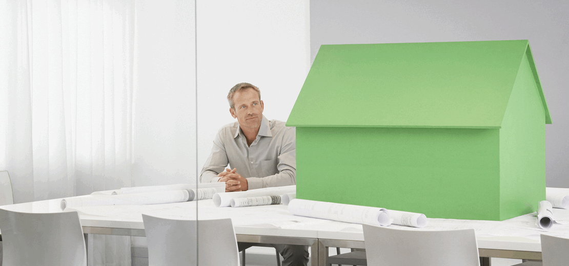 Mann sitzt an einem Tisch mit Bauplänen, daneben steht ein großes grünes Modellhaus. Das Motiv symbolisiert Planung und Umsetzung energieeffizienter bzw. klimafreundlicher Gebäude sowie die Umweltprüfung beim Bebauungsplan.