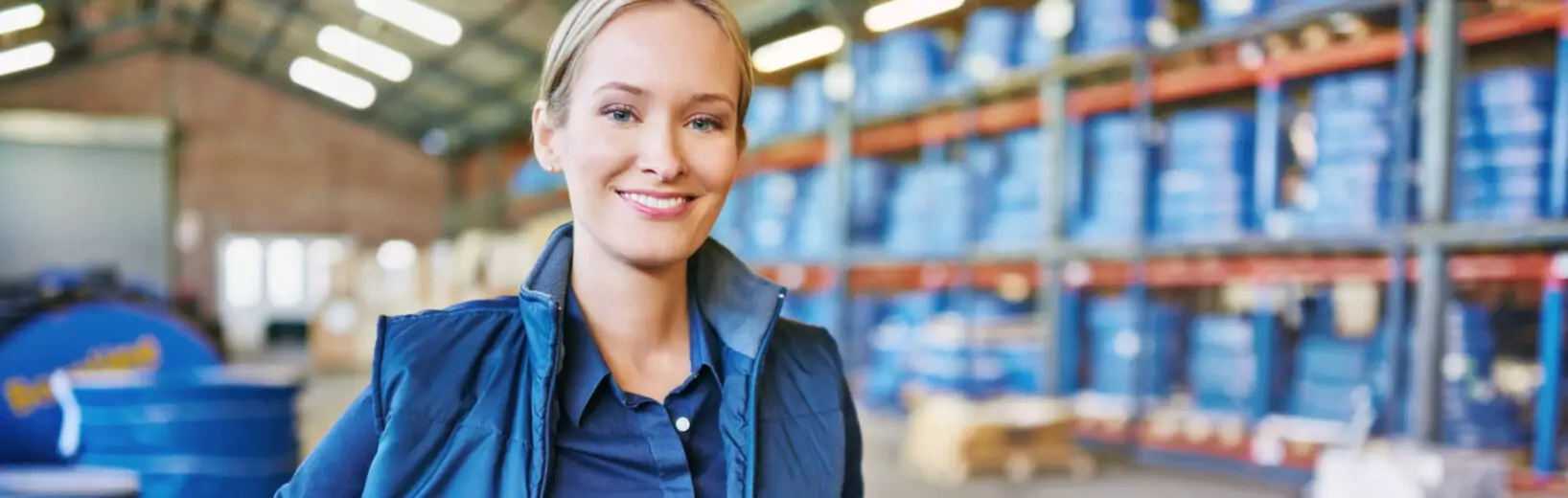 Frau lächelt in die Kamera, im Hintergrund Gefahrstofffässer. Symbolbild für Gefahrstoffbeauftragter.