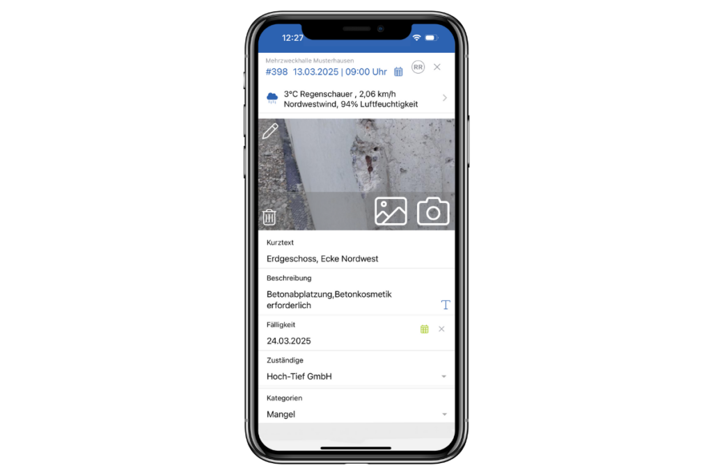 Auf dem Smartphone-Bildschirm wird eine Bau-App angezeigt, die einen konkreten Schaden detailliert beschreibt und die Baudokumentation „Vorfall Mangel“ integriert. Oben werden Wetterinformationen über einem Nahaufnahmefoto des Schadens angezeigt. Details umfassen Ort, Beschreibung, Priorität, Datum und das verantwortliche Unternehmen.