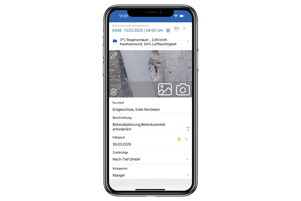 Auf dem Smartphone-Bildschirm wird eine Bau-App angezeigt, die einen konkreten Schaden detailliert beschreibt und die Baudokumentation „Vorfall Mangel“ integriert. Oben werden Wetterinformationen über einem Nahaufnahmefoto des Schadens angezeigt. Details umfassen Ort, Beschreibung, Priorität, Datum und das verantwortliche Unternehmen.