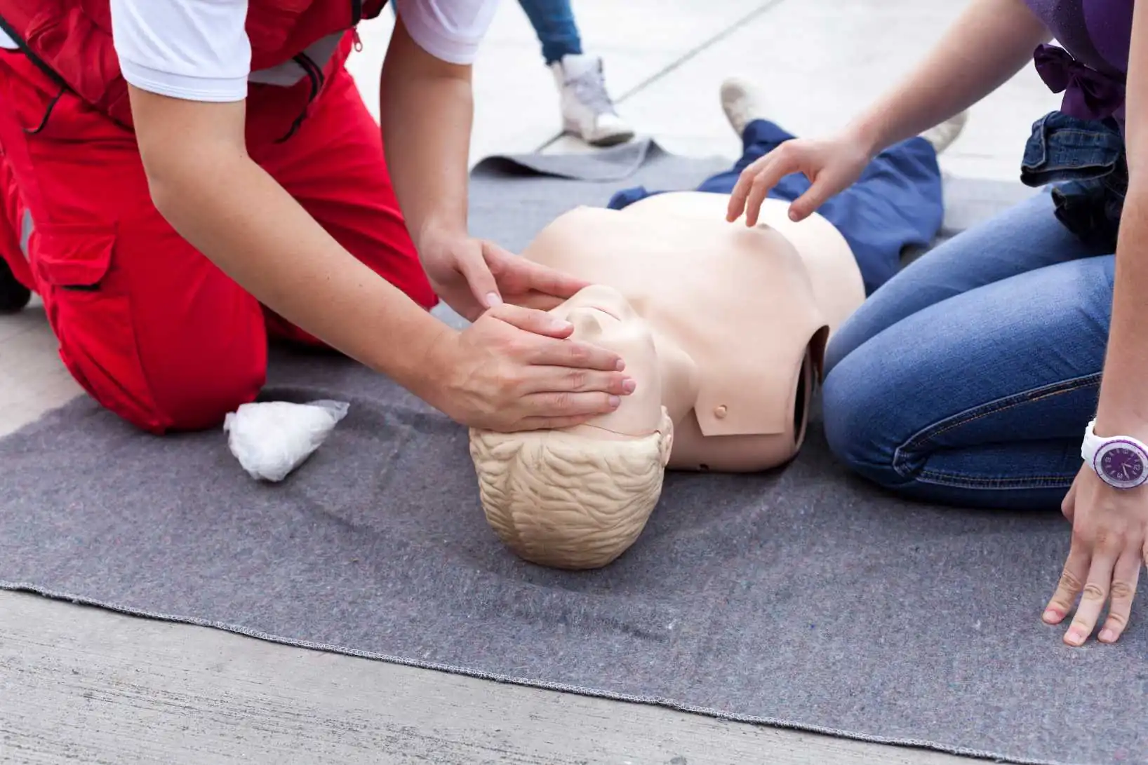 Zwei Personen knien neben einer Puppe auf dem Boden. Die Person links im Bild macht gerade Mund-zu-Mund-Beatmung bei der Puppe. Die Person rechts im Bild sucht gerade den Punkt für die Herzdruckmassage auf dem Oberkörper der Puppe. Das Bild zeigt eine typische Übung bei einem Erste-Hilfe-Lehrgang.
