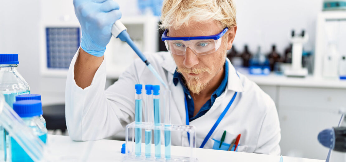 Mann in weißem Kittel im Labor betrachtet Reagenzgläschen mit blauer Flüssigkeit: Symbolbild für die Substitutionsprüfung.