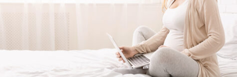 Junge schwangere Frau sitzt auf Bett und schaut in Laptop