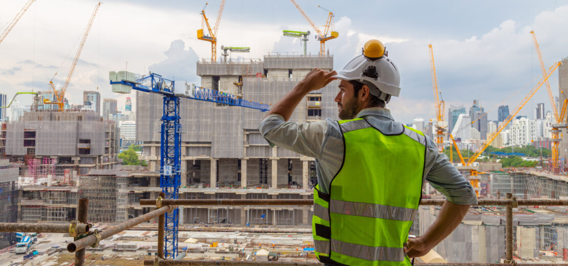 Ein Bauarbeiter in einer neongrünen Sicherheitsweste und einem weißen Helm überblickt eine sehr große Baustelle, auf der mehrere Kräne, unfertige Gebäude und die Skyline der Stadt im Hintergrund zu sehen sind. Das Bild vermittelt den Eindruck, dass viele Bauvorhaben gleichzeitig realisiert werden und an jeder Ecke gebaut wird. Dies untermautert den von der Regierung bezeichneteten "BauTurbo §246e", ein Gesetz zur Beschleunigung des Wohnungsbaus und zur Wohnraumsicherung.