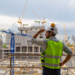 Ein Bauarbeiter in einer neongrünen Sicherheitsweste und einem weißen Helm überblickt eine sehr große Baustelle, auf der mehrere Kräne, unfertige Gebäude und die Skyline der Stadt im Hintergrund zu sehen sind. Das Bild vermittelt den Eindruck, dass viele Bauvorhaben gleichzeitig realisiert werden und an jeder Ecke gebaut wird. Dies untermautert den von der Regierung bezeichneteten "BauTurbo §246e", ein Gesetz zur Beschleunigung des Wohnungsbaus und zur Wohnraumsicherung.
