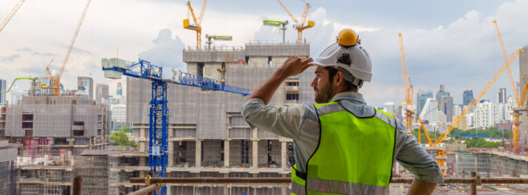 Ein Bauarbeiter in einer neongrünen Sicherheitsweste und einem weißen Helm überblickt eine sehr große Baustelle, auf der mehrere Kräne, unfertige Gebäude und die Skyline der Stadt im Hintergrund zu sehen sind. Das Bild vermittelt den Eindruck, dass viele Bauvorhaben gleichzeitig realisiert werden und an jeder Ecke gebaut wird. Dies untermautert den von der Regierung bezeichneteten "BauTurbo §246e", ein Gesetz zur Beschleunigung des Wohnungsbaus und zur Wohnraumsicherung.