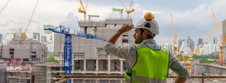 Ein Bauarbeiter in einer neongrünen Sicherheitsweste und einem weißen Helm überblickt eine sehr große Baustelle, auf der mehrere Kräne, unfertige Gebäude und die Skyline der Stadt im Hintergrund zu sehen sind. Das Bild vermittelt den Eindruck, dass viele Bauvorhaben gleichzeitig realisiert werden und an jeder Ecke gebaut wird. Dies untermautert den von der Regierung bezeichneteten "BauTurbo §246e", ein Gesetz zur Beschleunigung des Wohnungsbaus und zur Wohnraumsicherung.