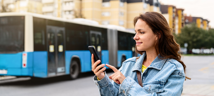 Frau benutzt ÖPNV-App am Smartphone, im Hinterrgund fährt ein Stadt-Linienbus