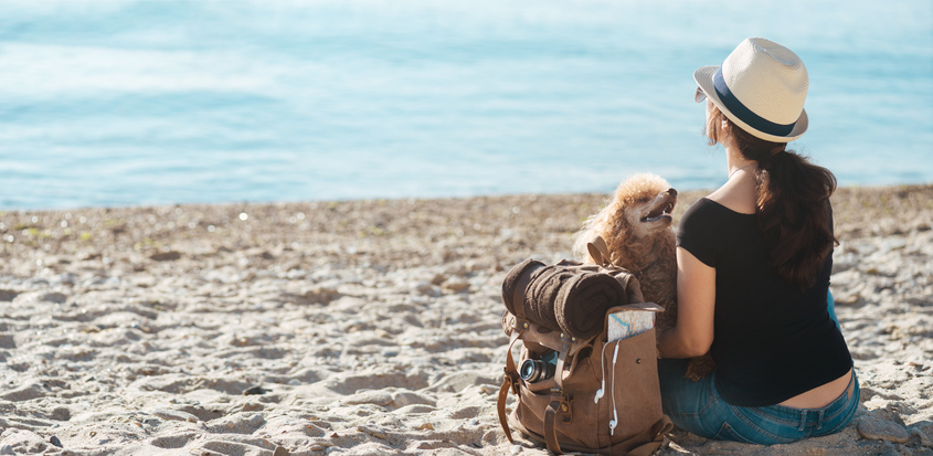 Ablehnung oder Streichung von Urlaub Junge Frau sitzt mit Pudel und Rucksack am Strand und genießt die Aussicht
