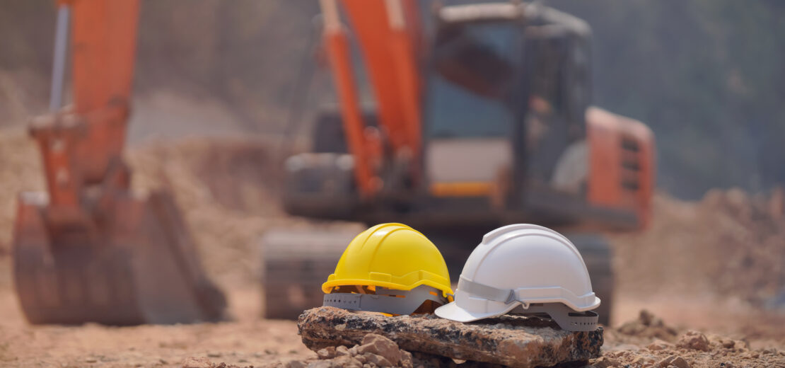 Zwei Bauhelme, ein gelber und ein weißer, ruhen auf einem Felsen an einer staubigen Erdarbeiten Baustelle. Im unscharfen Hintergrund ist ein orangefarbener Bagger zu sehen.