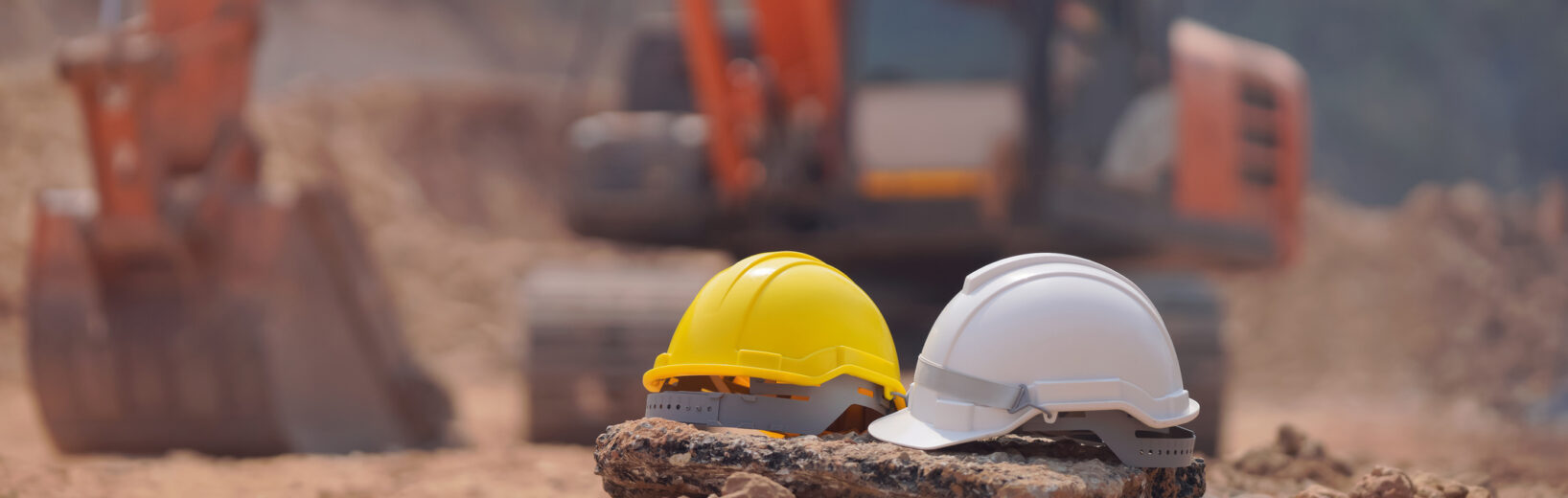 Allgemeine technische Vertragsbedingungen (ATV) – Geltungsbereich einfach erklärt Zwei Bauhelme, ein gelber und ein weißer, ruhen auf einem Felsen an einer staubigen Erdarbeiten Baustelle. Im unscharfen Hintergrund ist ein orangefarbener Bagger zu sehen.