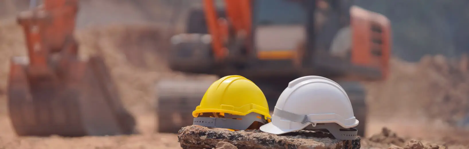 Zwei Bauhelme, ein gelber und ein weißer, ruhen auf einem Felsen an einer staubigen Erdarbeiten Baustelle. Im unscharfen Hintergrund ist ein orangefarbener Bagger zu sehen.