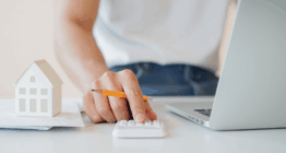 Eine Person, die in der Baubranche tätig ist, benutzt einen Taschenrechner an einem Schreibtisch mit einem Laptop und einem kleinen Modellhaus, das den Vorgang der Abrechnung nach VOB verdeutlicht; der Text Abrechnung nach VOB erscheint auf blauem Hintergrund.