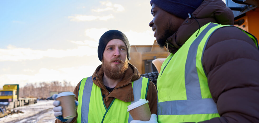 Portrait von zwei Bauarbeitern, die Kaffee trinken, um sich aufzuwärmen auf einer winterlichen Baustelle direkt neben einem schweren Flurförderzeug.