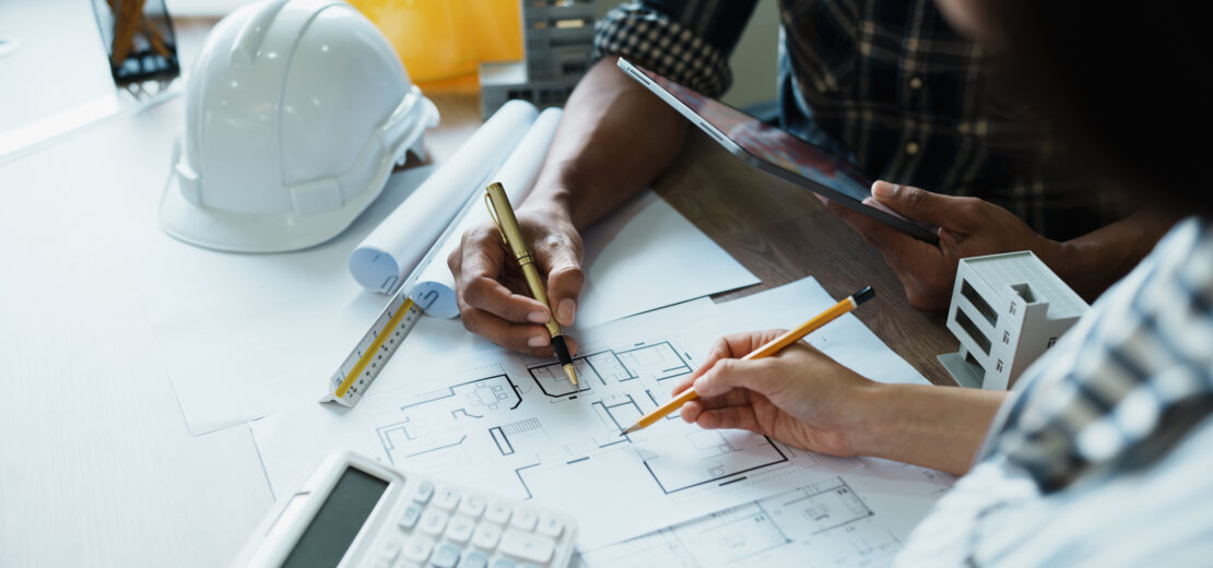 Zwei Personen arbeiten an einem Schreibtisch mit Bleistiften, einem Taschenrechner und einem Tablet an Architekturplänen. Auf dem Tisch liegen Bauwerkzeuge, ein weißer Schutzhelm und Unterlagen zur Besprechung der Nebenleistungen VOB