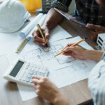 Zwei Personen arbeiten an einem Schreibtisch mit Bleistiften, einem Taschenrechner und einem Tablet an Architekturplänen. Auf dem Tisch liegen Bauwerkzeuge, ein weißer Schutzhelm und Unterlagen zur Besprechung der Nebenleistungen VOB