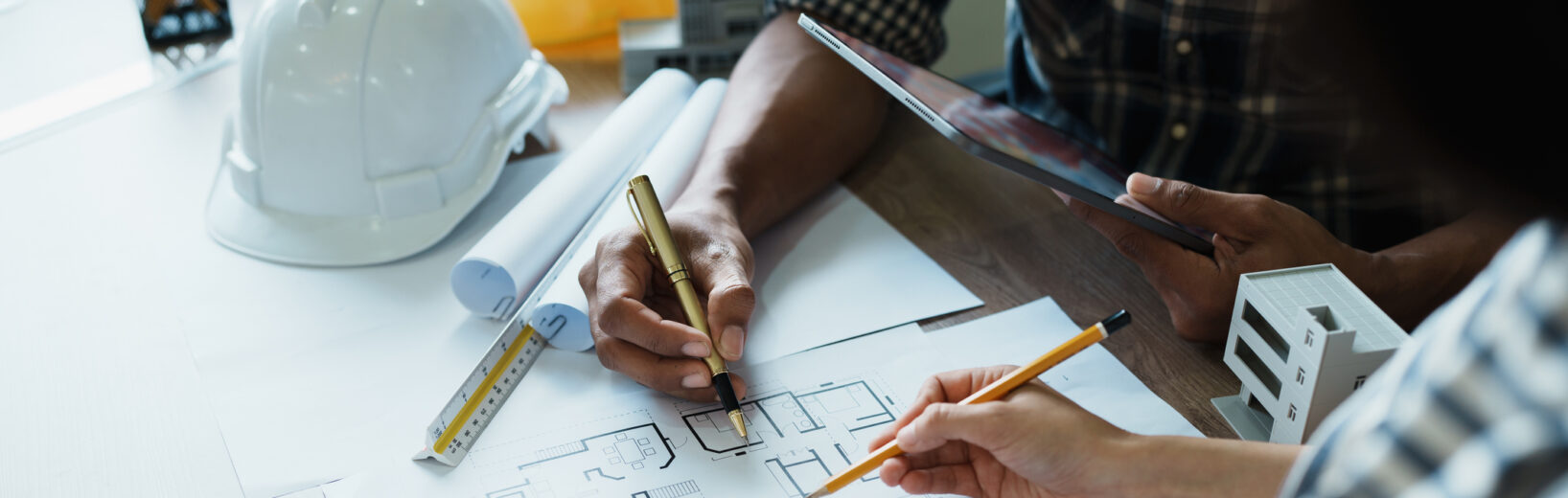 Zwei Personen arbeiten an einem Schreibtisch mit Bleistiften, einem Taschenrechner und einem Tablet an Architekturplänen. Auf dem Tisch liegen Bauwerkzeuge, ein weißer Schutzhelm und Unterlagen zur Besprechung der Nebenleistungen VOB