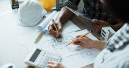 Zwei Personen arbeiten an einem Schreibtisch mit Bleistiften, einem Taschenrechner und einem Tablet an Architekturplänen. Auf dem Tisch liegen Bauwerkzeuge, ein weißer Schutzhelm und Unterlagen zur Besprechung der Nebenleistungen VOB