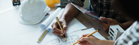 Zwei Personen arbeiten an einem Schreibtisch mit Bleistiften, einem Taschenrechner und einem Tablet an Architekturplänen. Auf dem Tisch liegen Bauwerkzeuge, ein weißer Schutzhelm und Unterlagen zur Besprechung der Nebenleistungen VOB