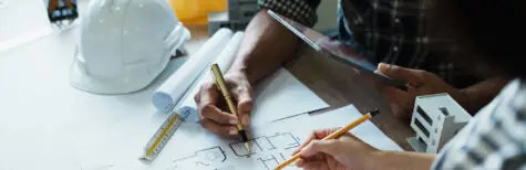 Zwei Personen arbeiten an einem Schreibtisch mit Bleistiften, einem Taschenrechner und einem Tablet an Architekturplänen. Auf dem Tisch liegen Bauwerkzeuge, ein weißer Schutzhelm und Unterlagen zur Besprechung der Nebenleistungen VOB