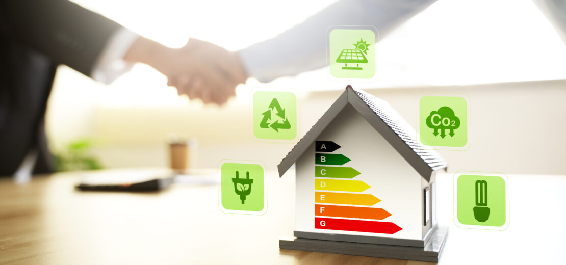 Modellhaus mit Energieeffizienzskala von A bis G, umgeben von Symbolen für Photovoltaik, CO₂-Reduktion, Recycling und Energieeffizienz. Im unscharfen Hintergrund reichen sich zwei Personen die Hand – als Symbol für nachhaltiges und energieeffizientes Bauen und Modernisieren.