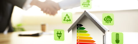 Modellhaus mit Energieeffizienzskala von A bis G, umgeben von Symbolen für Photovoltaik, CO₂-Reduktion, Recycling und Energieeffizienz. Im unscharfen Hintergrund reichen sich zwei Personen die Hand – als Symbol für nachhaltiges und energieeffizientes Bauen und Modernisieren.