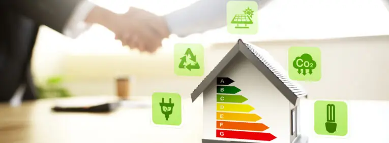 Modellhaus mit Energieeffizienzskala von A bis G, umgeben von Symbolen für Photovoltaik, CO₂-Reduktion, Recycling und Energieeffizienz. Im unscharfen Hintergrund reichen sich zwei Personen die Hand – als Symbol für nachhaltiges und energieeffizientes Bauen und Modernisieren.